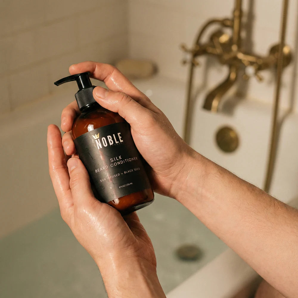 Hand holding a bottle of Noble Silk Beard Conditioner in a bathroom. Noble Product Silk Beard Conditioner texture – rich conditioning formula for beard softness and strength.