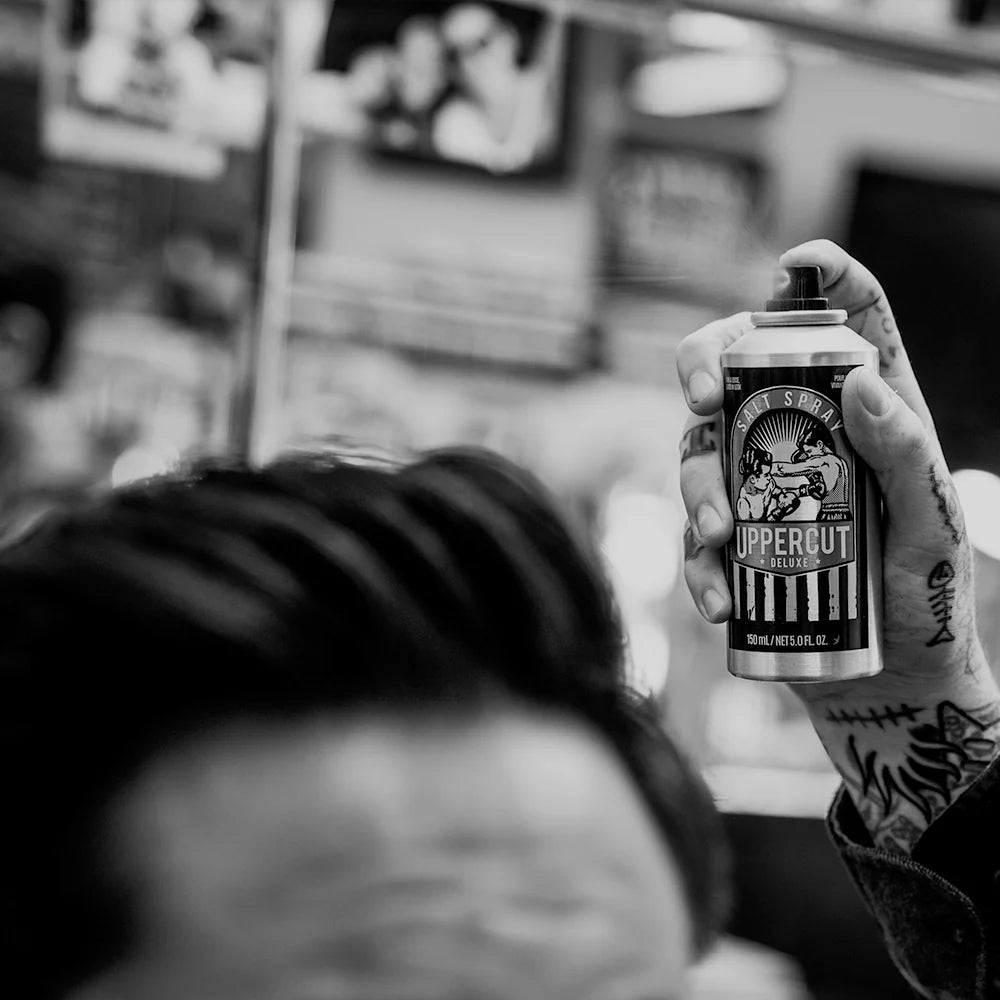 Man styling hair with Uppercut Salt Spray – loose textured lived-in look, light hold natural finish. Person holding a can of Uppercut Deluxe hair product with a blurred background